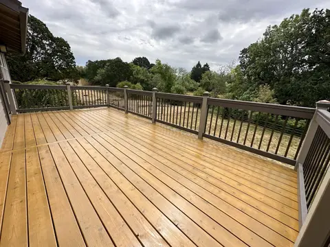 Composite deck with linear decking boards and black aluminum railing, surrounding mature trees under partly cloudy sky.