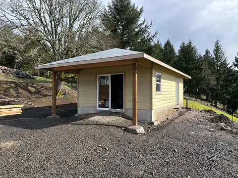 Finished ADU or small residential building with tan siding, metal roof, covered entry porch, and gravel pad, set on hillside