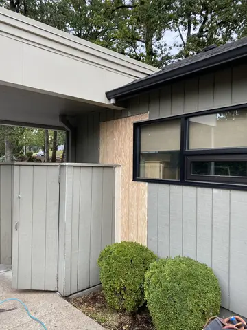 Modern home exterior featuring light gray vertical metal siding, black-framed windows, dark roof overhang, metal gate with