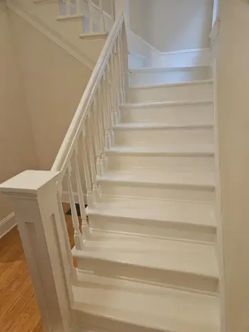 White painted staircase with white spindle railings descending to wood floor, light-filled stairwell with white walls.
