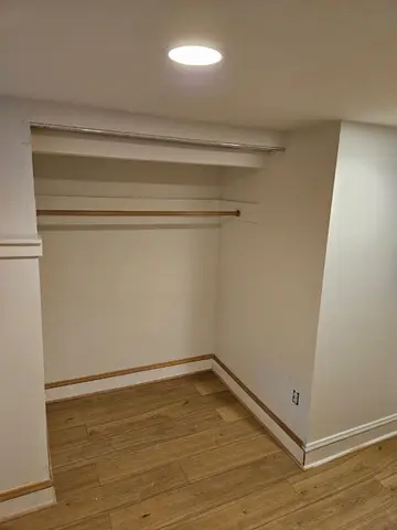 Empty walk-in closet with two wooden shelves, recessed ceiling light, hardwood flooring, and white walls.
