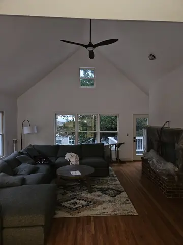 Bright living room with cathedral ceiling, black ceiling fan, large multi-pane windows overlooking trees, black sectional