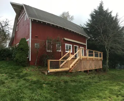 Red barn exterior with newly constructed wooden deck featuring stairs and metal railing, surrounded by evergreen trees and