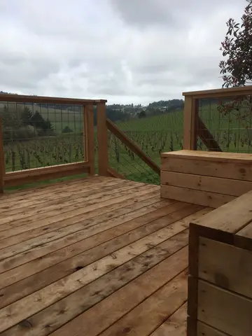 Wooden deck with horizontal planking and glass panel railings, overlooking green agricultural fields.