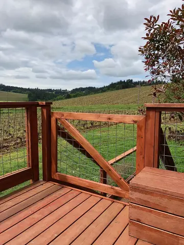 Redwood deck with metal-mesh gate and diagonal brace, overlooking rural farmland and evergreen forest under partly cloudy