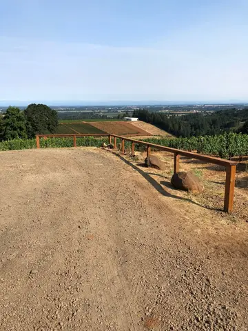 Wooden post-and-rail ranch fencing runs along a graded dirt driveway with forested hills and agricultural land visible in