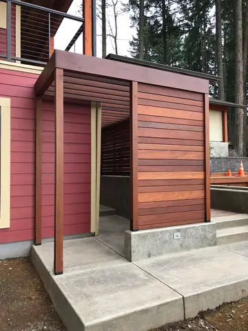Modern exterior structure with horizontal wood siding, steel frame posts, and open entry on concrete foundation pad.