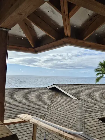 Newly shingled roof slope with ocean and sky visible beyond, framed by wooden beam structure and soffit.