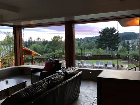 Modern living room with black leather sectional sofa facing large multi-panel windows, exterior view of forest, mountains