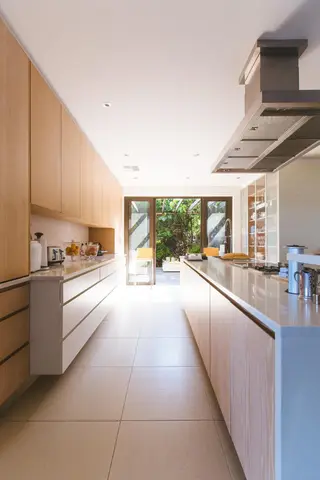 Bright galley kitchen with light wood cabinetry, white countertops, pale tile flooring, and glass doors opening to garden