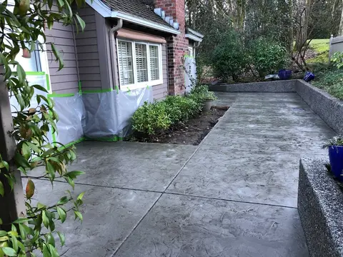 Finished concrete walkway with control joints running along side of house, bordered by landscaped planter beds and dark