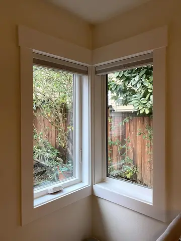 Two white-framed casement windows meeting at an interior corner with tan drywall and white trim, garden visible outside.