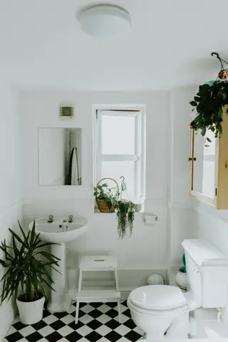 Bright bathroom with black-and-white checkered tile floor, white pedestal sink, toilet, and window with potted plants on