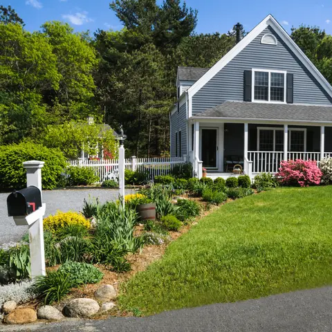 White farmhouse with gabled roof, front porch, mailbox, flowering shrubs, and sloped lawn with landscaped beds.