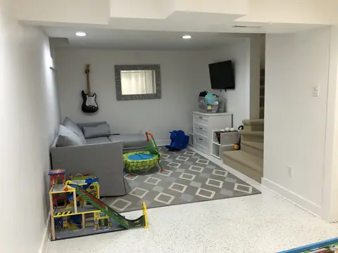 Finished basement playroom with gray geometric patterned tile flooring, gray upholstered seating along left wall, toy